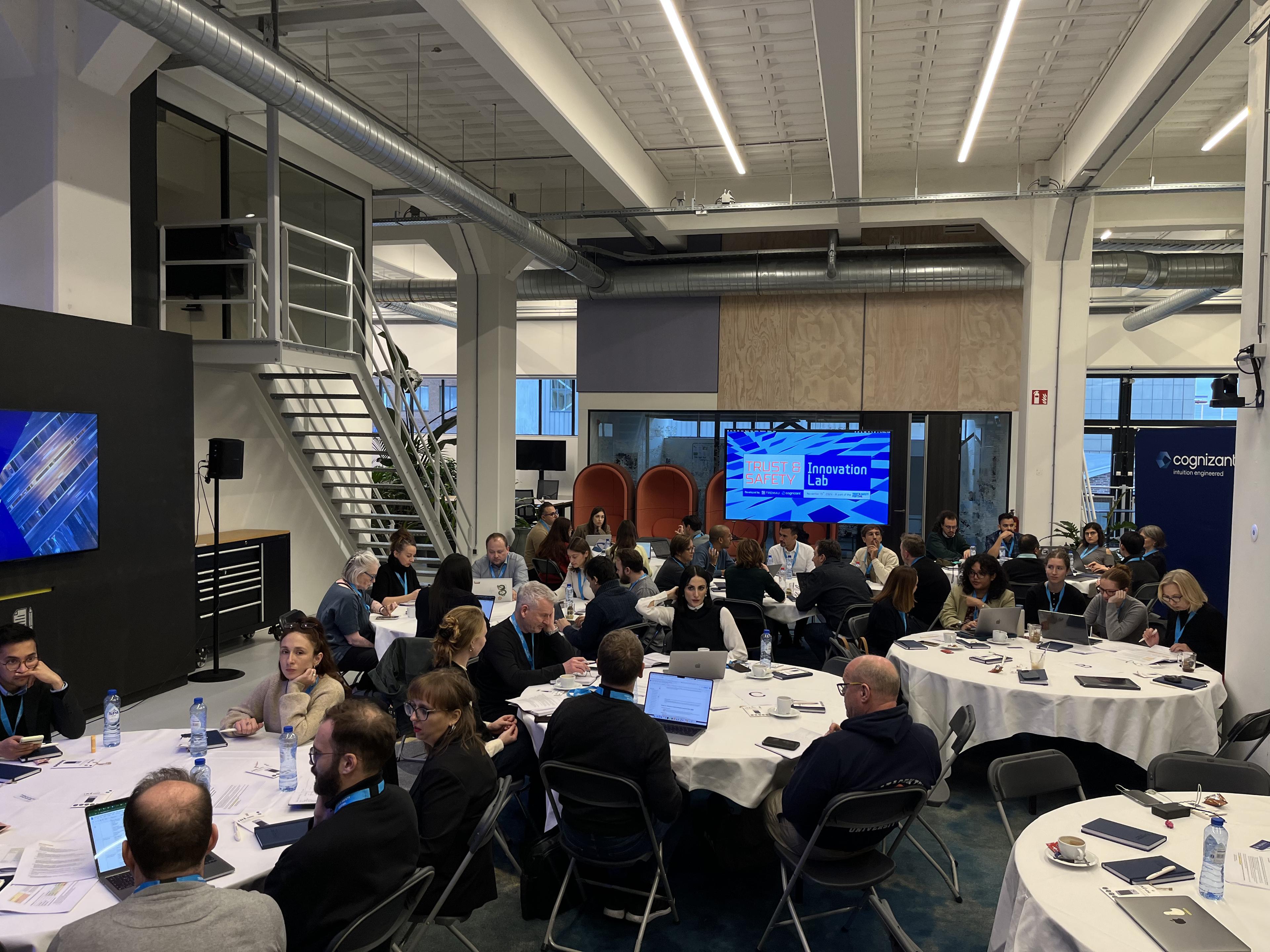 People collaborating around round tables during a Trust & Safety Innovation Lab workshop in a modern office space.