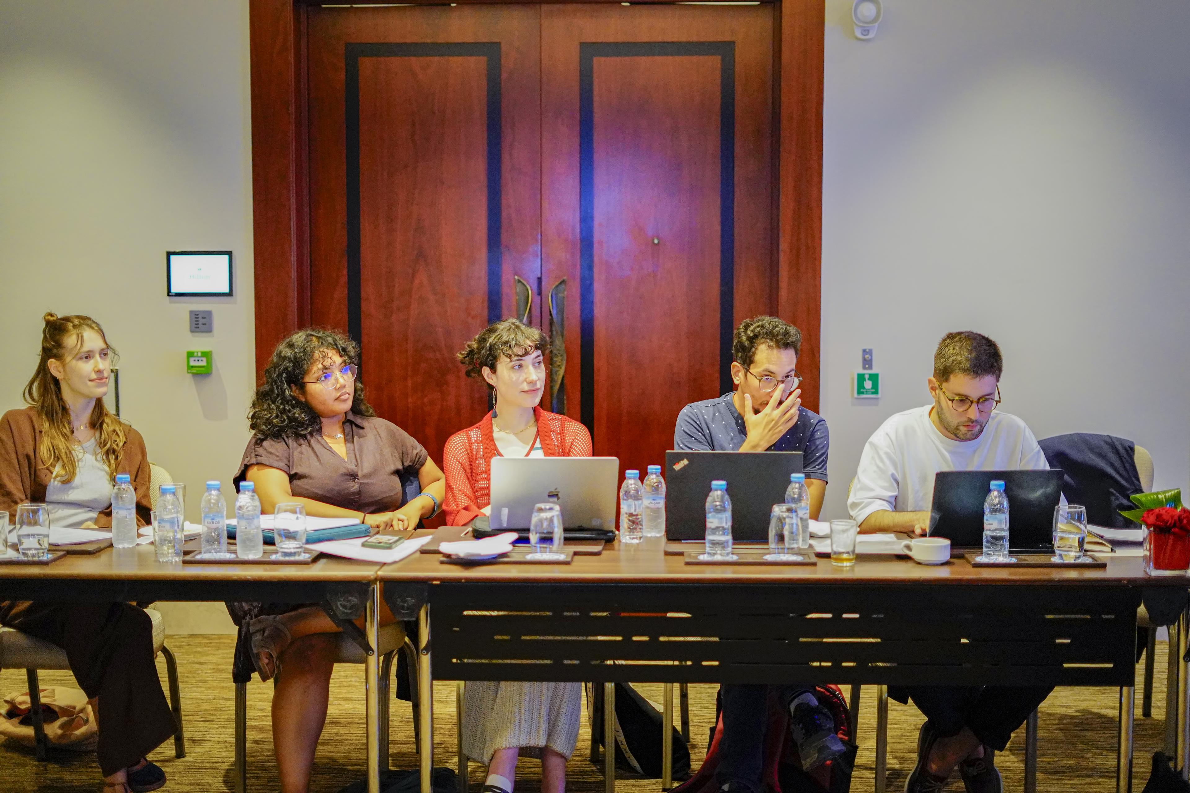 Panel discussion with people seated at a long table using laptops and notebooks.